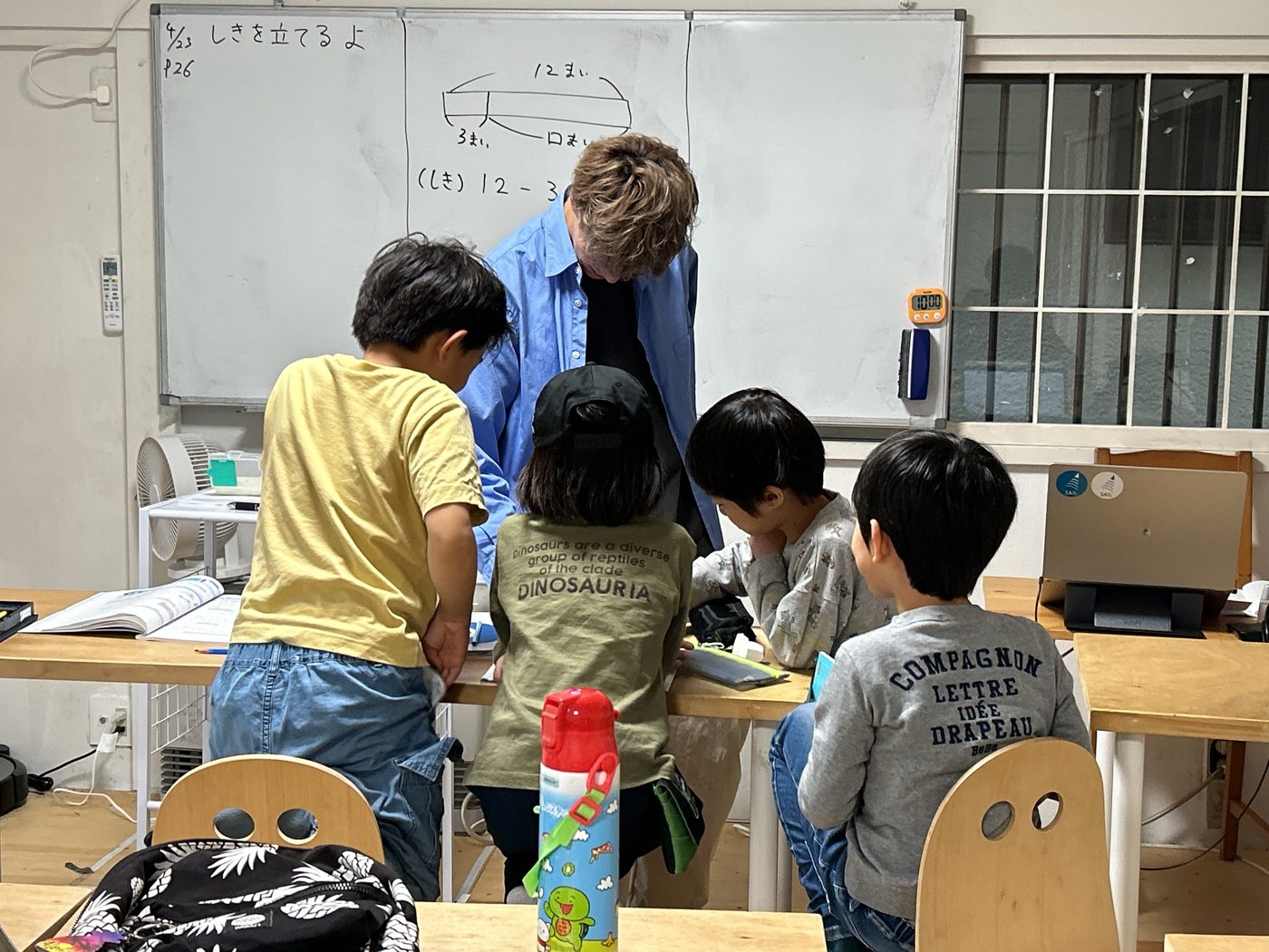 板書の前に立つ先生と、机に向かう小学生の後ろ姿